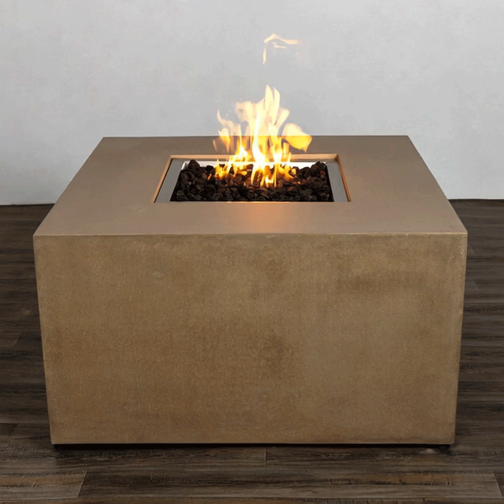 A sand-colored concrete fire pit in a simple studio setting takes the form of a low, solid blocky square fire pit, its smooth vertical sides resting on a dark wood plank floor, while the recessed central opening is packed with irregular black lava rocks and a bright yellow flame that rises in loose strands, the plain pale wall and clean geometry emphasizing a compact modern fire pit as the only object in the frame.