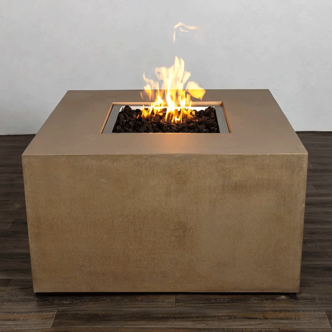 A sand-colored concrete fire pit in a simple studio setting takes the form of a low, solid blocky square fire pit, its smooth vertical sides resting on a dark wood plank floor, while the recessed central opening is packed with irregular black lava rocks and a bright yellow flame that rises in loose strands, the plain pale wall and clean geometry emphasizing a compact modern fire pit as the only object in the frame.