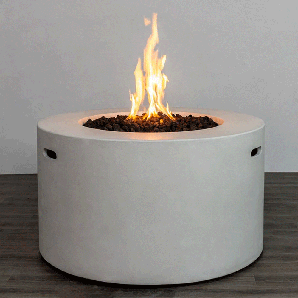 A bright, steady flame rises from a bed of dark lava rocks in a white round fire pit, the smooth cylindrical body of this concrete fire pit standing on a dark wood-look floor against a simple backdrop; the clean lines and integrated side handles highlight a sleek modern fire pit design, while the recessed burner of this gas fire pit delivers consistent, adjustable warmth for contemporary outdoor or semi-enclosed living spaces.