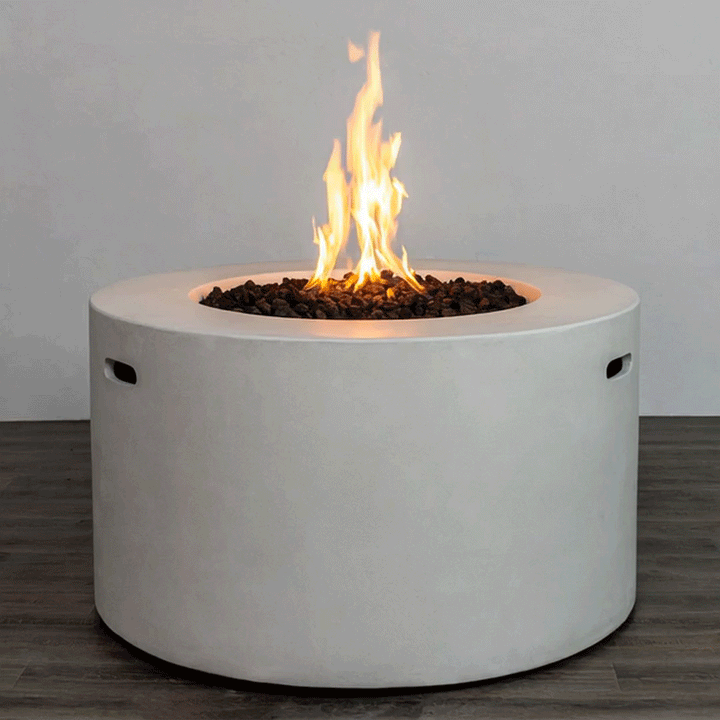 A bright, steady flame rises from a bed of dark lava rocks in a white round fire pit, the smooth cylindrical body of this concrete fire pit standing on a dark wood-look floor against a simple backdrop; the clean lines and integrated side handles highlight a sleek modern fire pit design, while the recessed burner of this gas fire pit delivers consistent, adjustable warmth for contemporary outdoor or semi-enclosed living spaces.