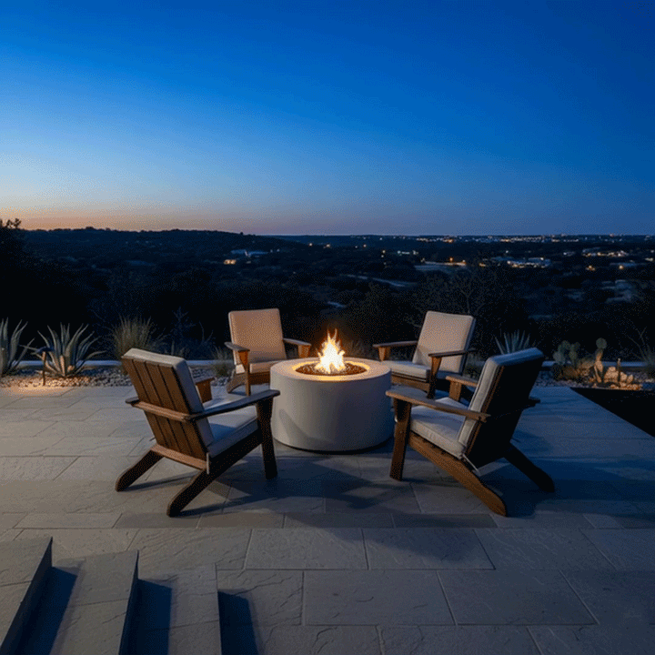At dusk on a raised stone terrace, four cushioned Adirondack chairs encircle a white round fire pit, its golden flames reflecting on the patio as the sky deepens to blue over distant hills and scattered lights below; this inviting patio fire pit serves as a central outdoor fire pit where friends or relatives can gather, talk, and roast snacks, functioning as a relaxed family gathering fire pit that extends evenings comfortably into the night.