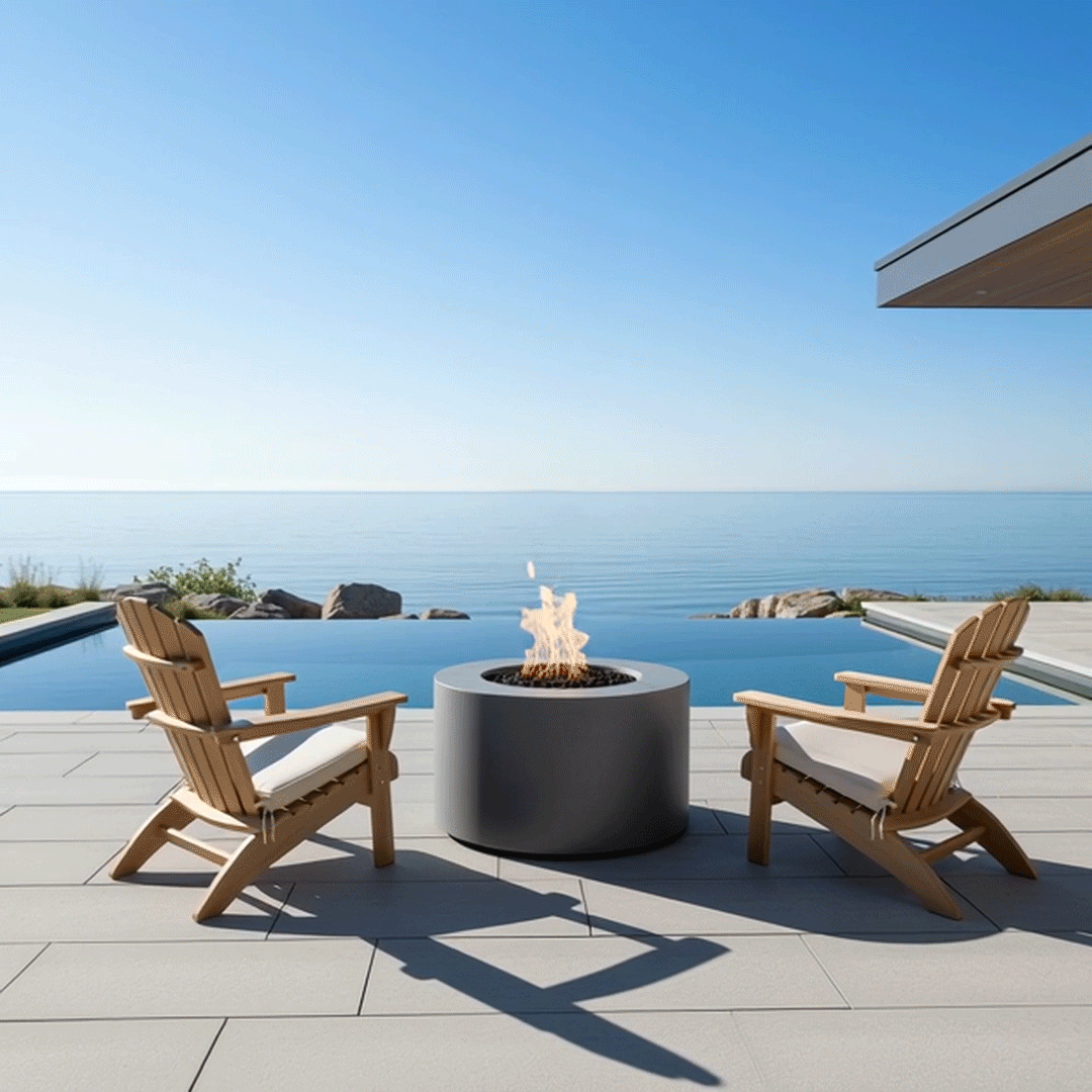 Overlooking a serene infinity pool that blends into a calm blue sea, two wooden Adirondack chairs face a compact gray round fire pit, its flames dancing gently above lava rocks on a broad stone terrace; this minimalist outdoor fire pit acts as a refined luxury fire pit centerpiece, transforming the waterfront patio into a peaceful backyard oasis where people can unwind after a swim, sip coffee at sunrise, or linger beside the water late into the evening.