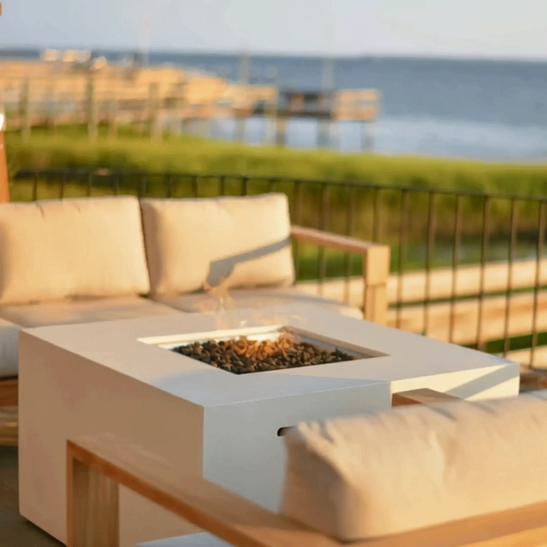 On a sunlit deck beside calm blue water and wooden docks, a low concrete fire pit forming a white square fire pit sits between pale cushioned wood-frame sofas, its central opening filled with rough dark lava rocks and a small yellow flame, while black railing bars, a strip of green grass, and the distant shoreline frame the relaxed scene, clearly placing this outdoor fire pit at the edge of a waterfront seating area.