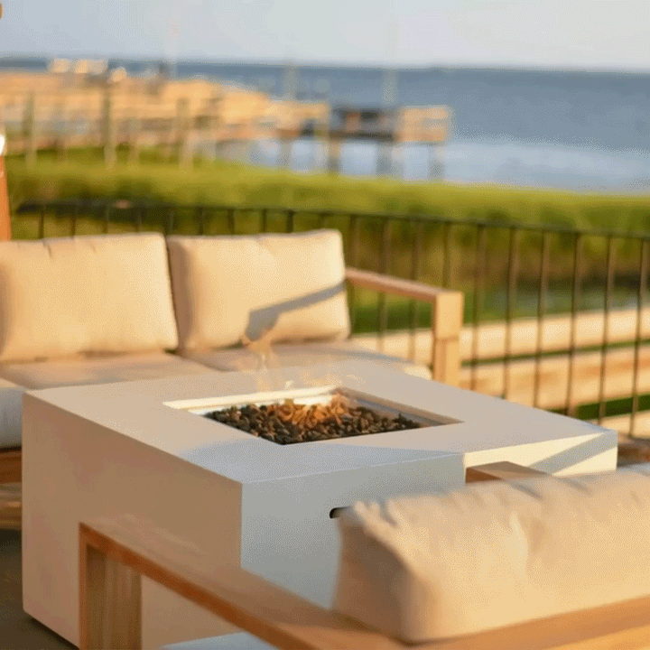 On a sunlit deck beside calm blue water and wooden docks, a low concrete fire pit forming a white square fire pit sits between pale cushioned wood-frame sofas, its central opening filled with rough dark lava rocks and a small yellow flame, while black railing bars, a strip of green grass, and the distant shoreline frame the relaxed scene, clearly placing this outdoor fire pit at the edge of a waterfront seating area.