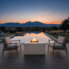 At dusk on a wide terrace facing an infinity pool and distant mountains, two cushioned armchairs with wooden arms sit on either side of a white patio fire pit, the smooth blocky shape revealing a concrete fire pit with a square fire pit opening in the center that holds dark stones and a compact flame, while trimmed grass, low plantings, and the reflected sky in the still water complete the quiet rectangular layout.