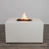 In a plain studio-style setting, a solid white concrete fire pit with straight sides stands on a dark wood floor, the piece shaped as a low cube with a centered square fire pit opening cut into the top surface, where jagged dark lava rocks fill the recess and a steady orange flame rises, the clean edges, smooth finish, and lack of additional objects emphasizing a simple modern fire pit presented alone against a light wall.