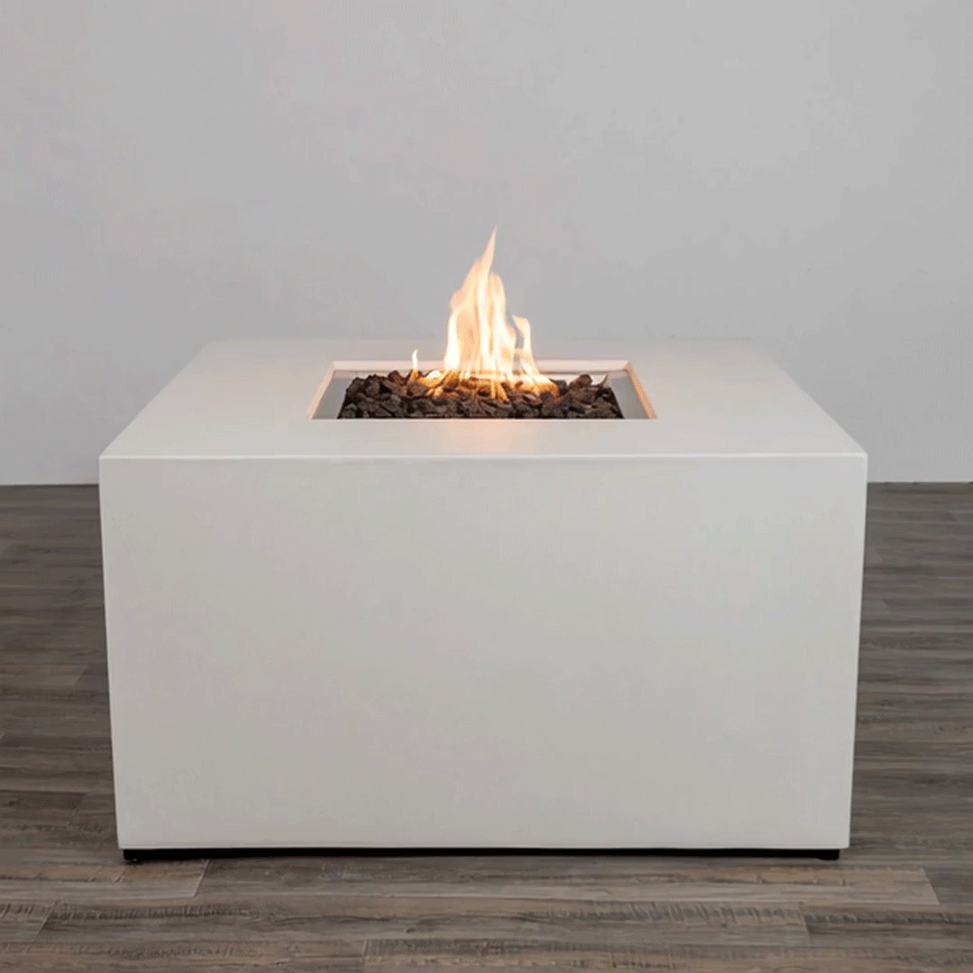 In a plain studio-style setting, a solid white concrete fire pit with straight sides stands on a dark wood floor, the piece shaped as a low cube with a centered square fire pit opening cut into the top surface, where jagged dark lava rocks fill the recess and a steady orange flame rises, the clean edges, smooth finish, and lack of additional objects emphasizing a simple modern fire pit presented alone against a light wall.