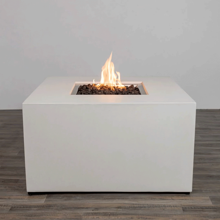 In a plain studio-style setting, a solid white concrete fire pit with straight sides stands on a dark wood floor, the piece shaped as a low cube with a centered square fire pit opening cut into the top surface, where jagged dark lava rocks fill the recess and a steady orange flame rises, the clean edges, smooth finish, and lack of additional objects emphasizing a simple modern fire pit presented alone against a light wall.