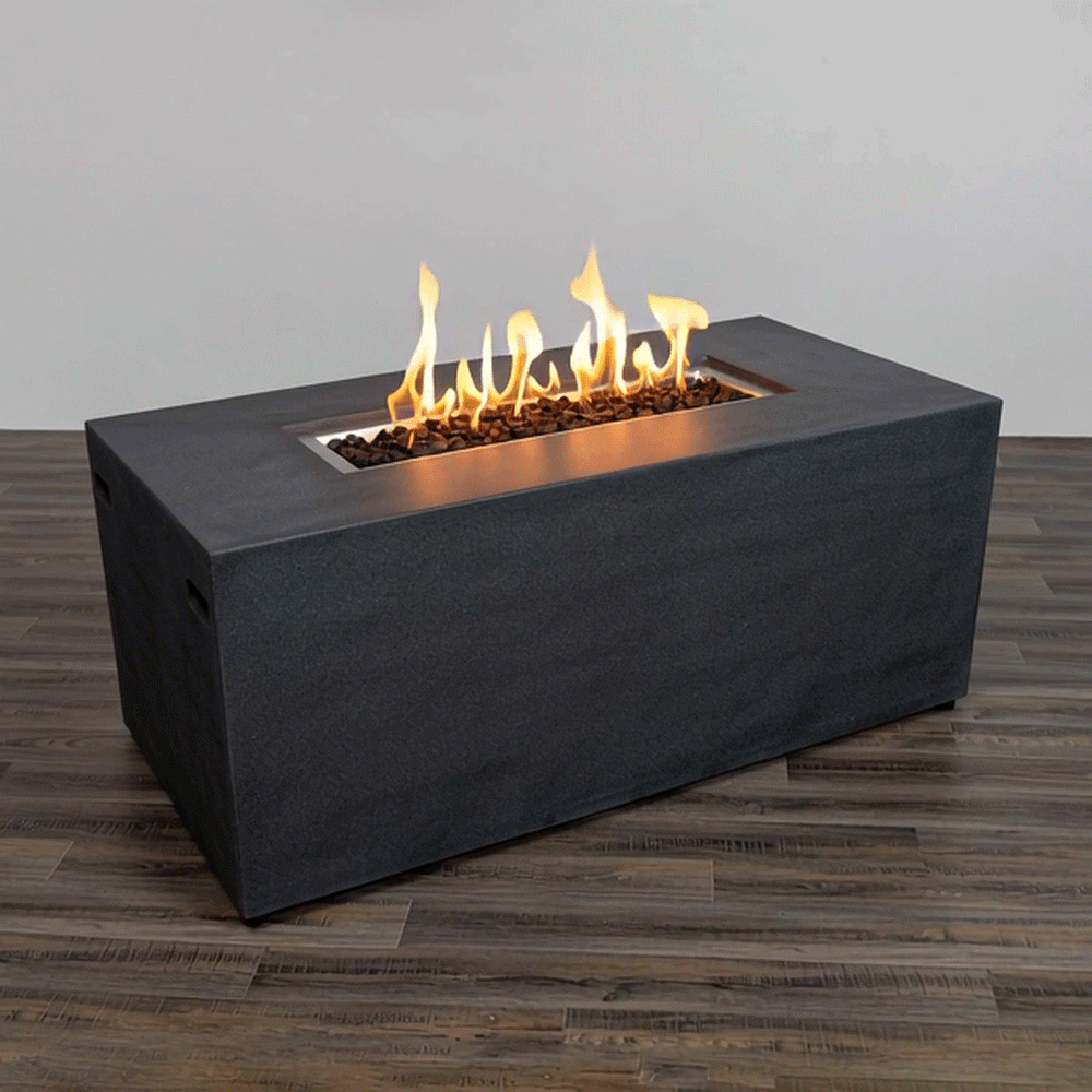 A charcoal gray rectangular fire pit with a low, boxy shape sits on a dark wood plank floor against a plain pale wall, the smooth surface and straight edges giving the concrete fire pit a minimalist modern fire pit look while a long recessed opening stretches along the top center, filled with rough dark lava rocks from which a line of bright yellow flames rises in uneven, flickering peaks.