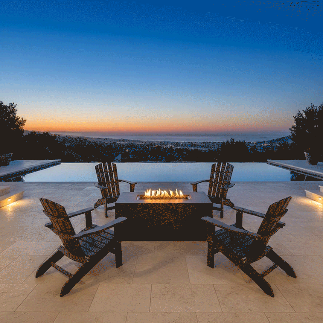 On a light tiled terrace overlooking coastal rooftops and the ocean at sunset, two cushioned armchairs face a charcoal rectangular fire pit placed near the edge of an infinity pool, the low outdoor fire pit showing a narrow central opening packed with dark stones and a row of small flames, flanked by square planters of spiky greenery, the patio fire pit positioned so the seating arrangement suggests relaxed outdoor entertaining above the shoreline.