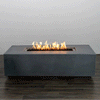 In a plain studio setting, a long low rectangular fire pit made from smooth natural gray concrete sits on a dark wood floor, its simple block shape giving the appearance of a modern fire pit, while a narrow recessed opening runs along the top center, packed with rough dark lava rocks and a continuous row of yellow flames that flicker upward against a pale wall.