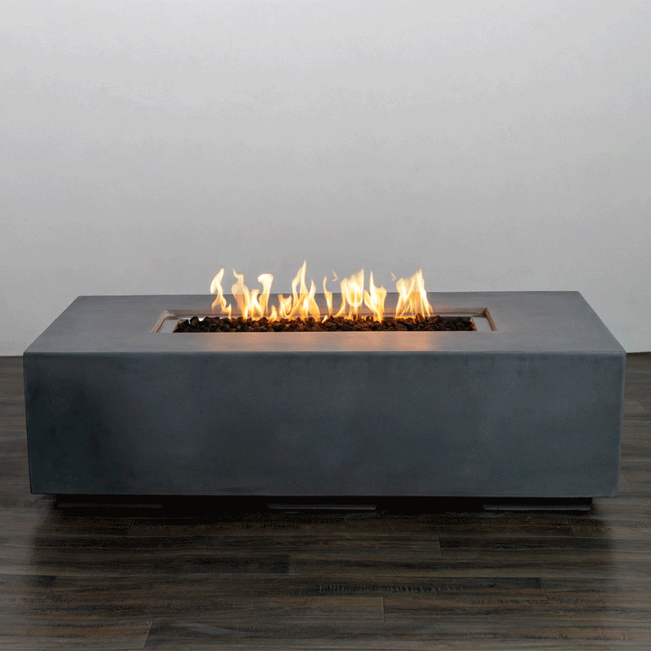 In a plain studio setting, a long low rectangular fire pit made from smooth natural gray concrete sits on a dark wood floor, its simple block shape giving the appearance of a modern fire pit, while a narrow recessed opening runs along the top center, packed with rough dark lava rocks and a continuous row of yellow flames that flicker upward against a pale wall.