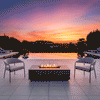 On a tiled terrace beside an infinity pool at sunset, two slim metal lounge chairs face a charcoal rectangular fire pit positioned near the water’s edge, the patio fire pit showing a linear channel of flames above dark stones, with the pool reflecting the orange and purple sky and low hills in the distance, so the glowing outdoor fire pit becomes the central fire feature in the open seating area.