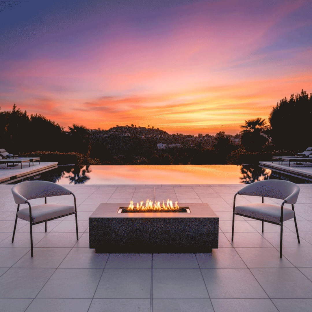 On a tiled terrace beside an infinity pool at sunset, two slim metal lounge chairs face a charcoal rectangular fire pit positioned near the water’s edge, the patio fire pit showing a linear channel of flames above dark stones, with the pool reflecting the orange and purple sky and low hills in the distance, so the glowing outdoor fire pit becomes the central fire feature in the open seating area.