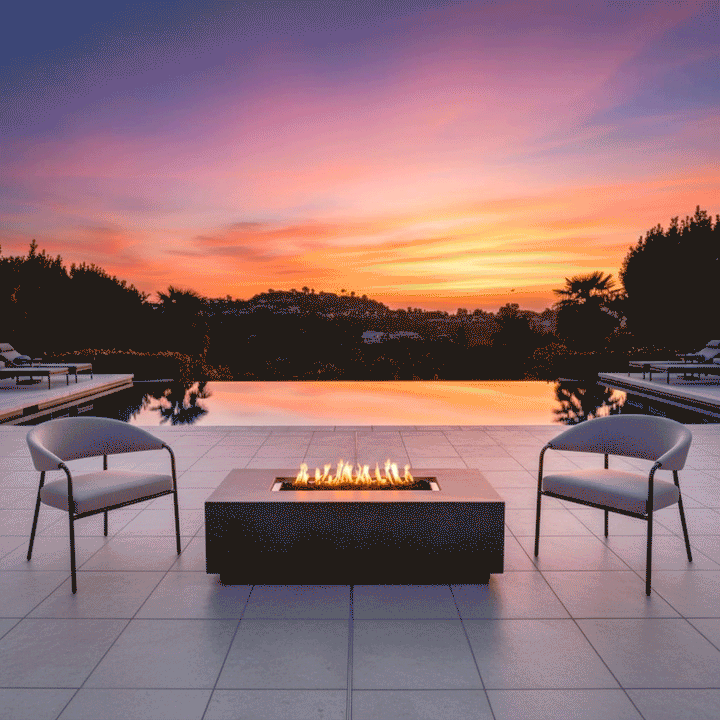 On a tiled terrace beside an infinity pool at sunset, two slim metal lounge chairs face a charcoal rectangular fire pit positioned near the water’s edge, the patio fire pit showing a linear channel of flames above dark stones, with the pool reflecting the orange and purple sky and low hills in the distance, so the glowing outdoor fire pit becomes the central fire feature in the open seating area.