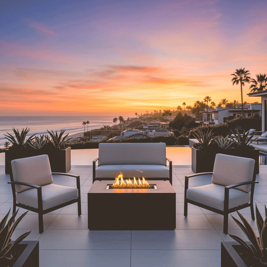 On a tiled terrace above the ocean at sunset, a low charcoal rectangular fire pit made of smooth concrete sits between a cushioned loveseat and two matching lounge chairs, its long central opening filled with dark stones and a row of yellow flames, surrounded by square planters of succulents and framed by palm trees, rooftops, and shoreline, arranged as an outdoor fire pit grouping suitable for relaxed outdoor entertaining with a wide coastal view.