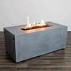 In a simple studio setting, a natural gray rectangular fire pit with straight sides and subtle handholds rests on a dark wood plank floor against a pale wall, the smooth surface identifying it as a concrete fire pit while a long recessed opening runs along the top center, holding uneven black lava rocks and a continuous row of bright flames, the clean geometry and lack of other objects emphasizing a minimal modern fire pit shape.