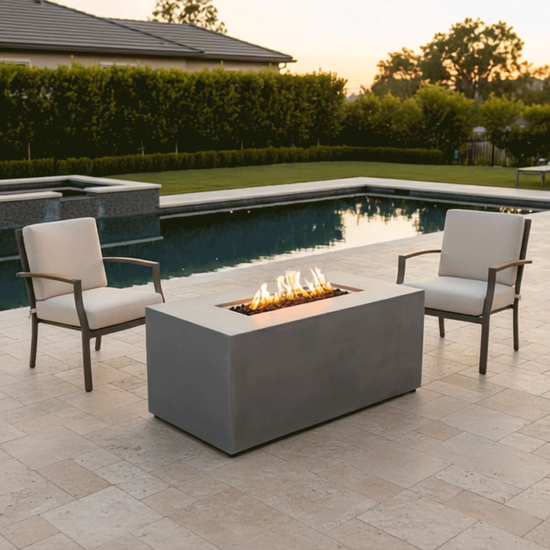 On a light stone patio overlooking a rectangular swimming pool at dusk, two upholstered armchairs with slim arms face each other across a gray rectangular fire pit, the low patio fire pit containing a narrow trough of dark lava rocks and a neat line of golden flames, with trimmed hedges, lawn, and distant trees forming the backdrop so the arrangement reads visually as a straightforward backyard fire pit seating area beside the water.