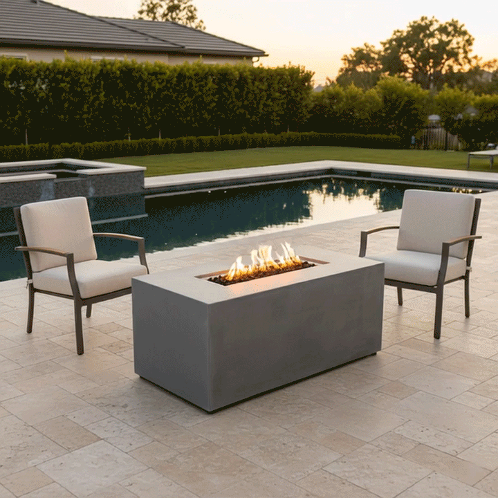 On a light stone patio overlooking a rectangular swimming pool at dusk, two upholstered armchairs with slim arms face each other across a gray rectangular fire pit, the low patio fire pit containing a narrow trough of dark lava rocks and a neat line of golden flames, with trimmed hedges, lawn, and distant trees forming the backdrop so the arrangement reads visually as a straightforward backyard fire pit seating area beside the water.