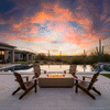 On a pale stone patio beside an infinity pool that blends into a desert horizon, four wooden Adirondack chairs form a circle around a low sand-colored concrete fire pit, its long rectangular burner sending steady flames above dark lava rocks as tall saguaro cacti and a vivid pink-and-orange sunset sky frame the scene, turning the space into a relaxed backyard fire pit setting that feels like a private backyard oasis in the desert.
