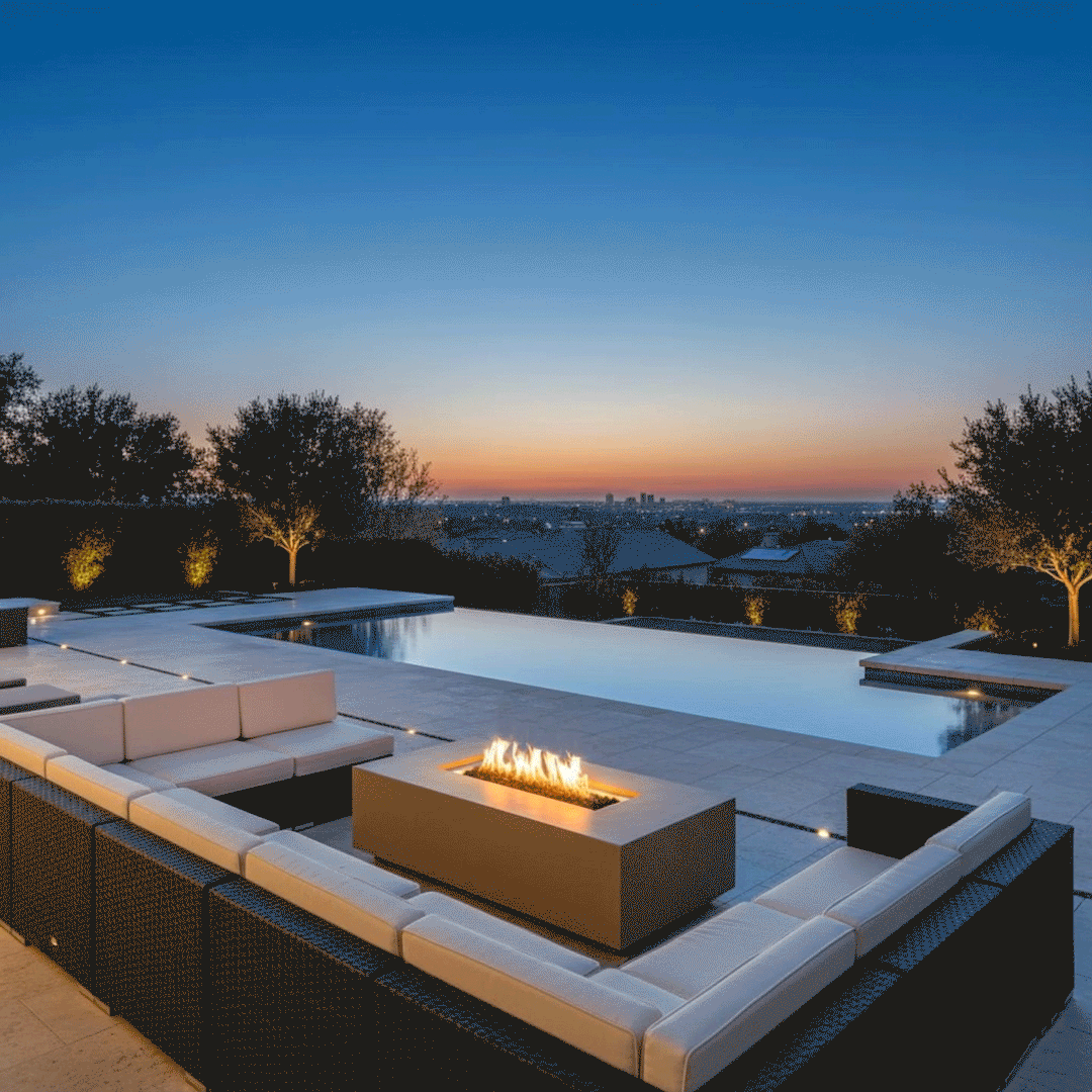 At dusk on a broad poolside terrace, a tan concrete fire pit with a slim rectangular opening and bright linear flames sits at the center of a U-shaped wicker sectional topped with light cushions, while an infinity pool reflects the fading orange skyline and softly lit trees, creating an inviting outdoor fire pit arrangement that supports casual conversation, lounging, and flexible outdoor entertaining with city views in the distance.