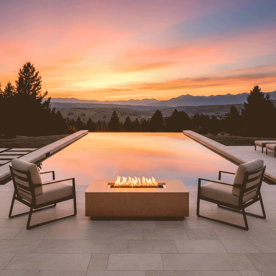 Two cushioned lounge chairs face each other across a low sand-colored concrete fire pit, its elongated rectangular burner casting warm light onto the stone patio as flames flicker over dark stones, all set at the edge of an infinity pool that mirrors a soft peach and lavender sunset over rolling hills and dark tree silhouettes, presenting a quiet corner where the luxury fire pit and water together shape a tranquil backyard oasis.