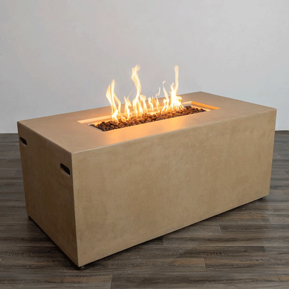 A sand-colored rectangular fire pit with clean, straight sides rests on a dark wood floor against a plain light wall, the smooth blocky body of this concrete fire pit framing a long recessed opening filled with uneven dark lava rocks, where a continuous row of yellow flames rises and bends slightly, the uncluttered studio setting emphasizing the low modern fire pit as the only object in view.
