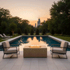On a light stone terrace beside a narrow pool at sunset, two metal-framed lounge chairs with beige cushions face a sand-toned patio fire pit shaped as a low rectangular fire pit, its central channel of flames reflecting in the still water while trees and a distant city skyline frame the yard, the simple seating around this backyard fire pit suggesting a compact outdoor entertaining area at the edge of the lawn.