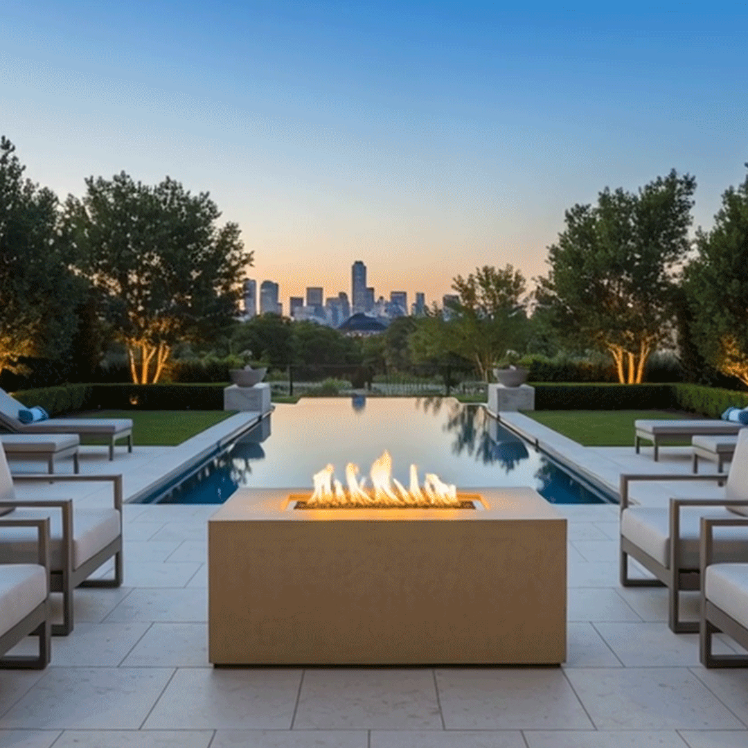 In a tree-lined yard at dusk, multiple cushioned chairs are arranged along a long pool facing a sand-colored rectangular fire pit, the outdoor fire pit set on pale tiles and showing a neat row of flames above dark stones, with manicured hedges, softly lit trees, and a faint skyline beyond, creating the look of a cozy backyard fire pit that could serve as a casual family gathering fire pit beside the water.