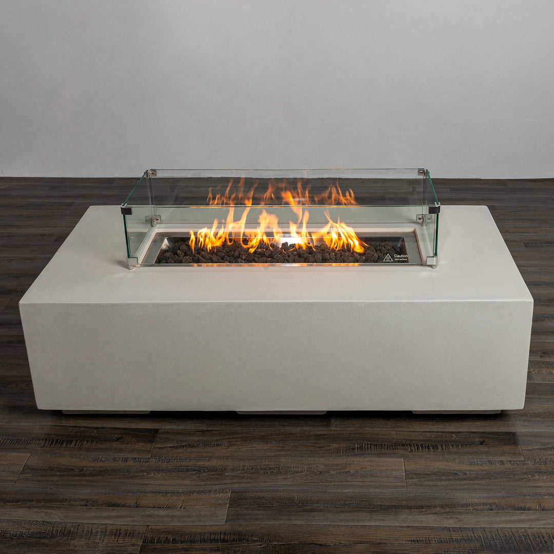 The Starfire Designs Beton 55" White Rectangle Gas Fire Pit lit with flames and protected by a glass wind guard. The brilliant flames dance over the black lava rocks, adding warmth and style to any outdoor space.