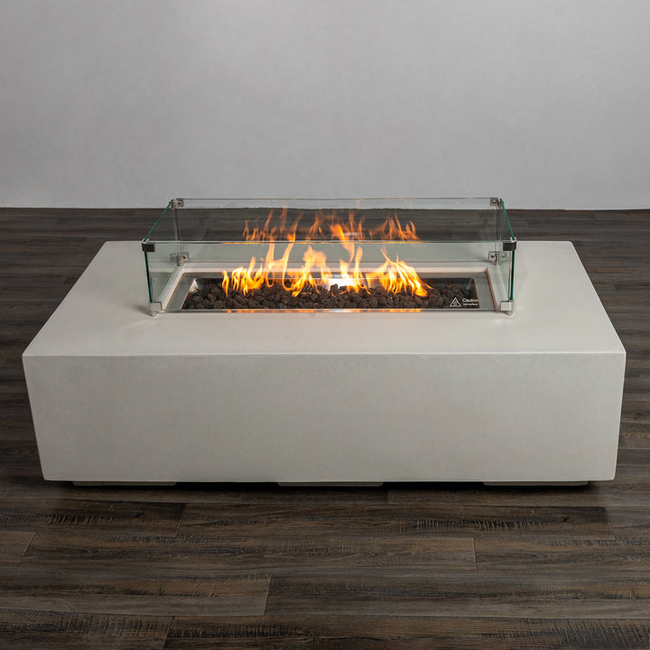 The Starfire Designs Beton 55" White Rectangle Gas Fire Pit lit with flames and protected by a glass wind guard. The brilliant flames dance over the black lava rocks, adding warmth and style to any outdoor space.