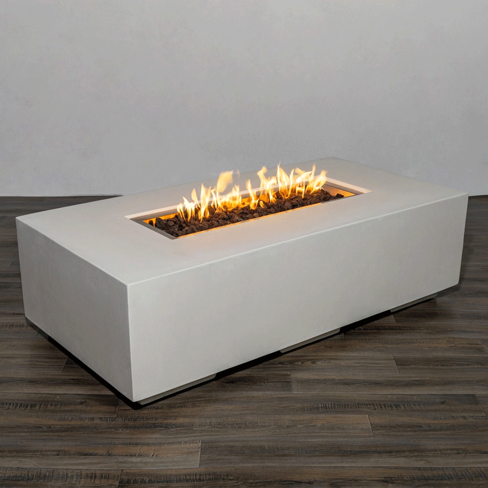 A low, modern concrete fire pit in a crisp white rectangular block sits alone on a warm wood-look floor against a plain gray wall, its long, narrow burner channel filled with chunky dark lava rocks while bright flames from the gas fire pit move in an even line, clearly revealing the sharp edges, smooth surface, and minimalist proportions of this substantial rectangular fire pit designed as a modern fire pit centerpiece for contemporary outdoor spaces and patios.