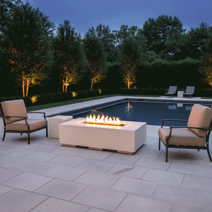 At twilight beside a rectangular backyard pool, a low white concrete fire pit glows at the center of three cushioned lounge chairs, its long linear flames flickering over dark lava rocks while trees and hedges lit from below frame the scene, presenting a cozy backyard fire pit layout where reflections dance on the still water and the modern fire pit naturally becomes the gathering spot for quiet outdoor entertaining, conversation, and stargazing late into the evening.