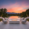 Framed by an infinity pool reflecting a vivid sunset and distant mountains, a low white concrete fire pit with a long, narrow flame channel anchors four matching lounge chairs on a broad stone terrace, creating a perfectly symmetrical backyard oasis view where the luxury fire pit reads as a sleek rectangular fire pit designed for refined yet comfortable outdoor relaxation, quiet conversation, and unhurried appreciation of the changing colors in the sky.