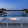 On a wide terrace overlooking a calm bay, two low modern sectionals face each other across a white rectangular fire pit placed near the edge of an infinity pool, the outdoor fire pit showing a continuous row of yellow flames above small stones while palm trees and distant buildings reflect in the still water; the linear fire feature and surrounding palms make the open seating area resemble a waterfront backyard oasis at blue hour.