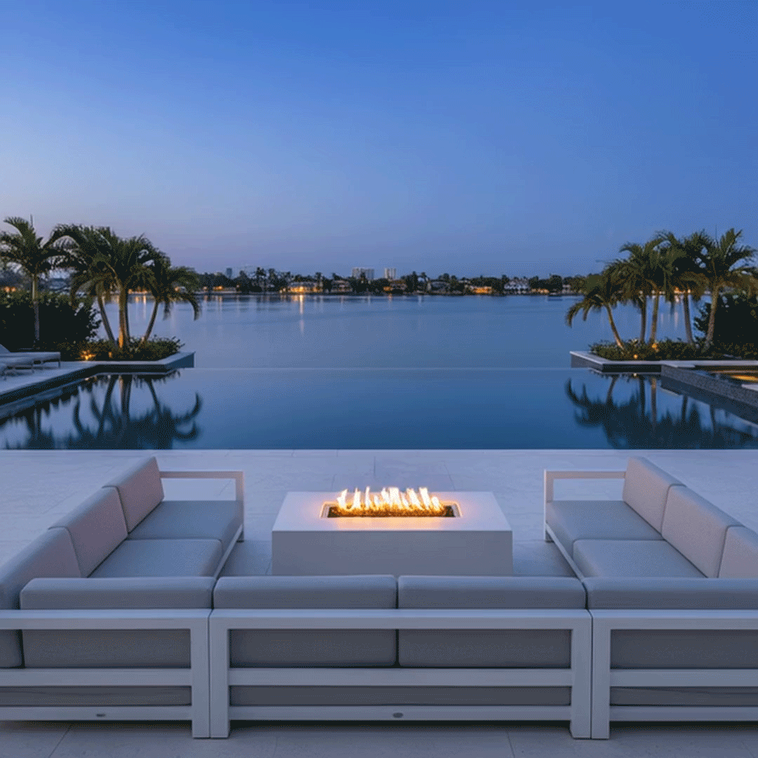 On a wide terrace overlooking a calm bay, two low modern sectionals face each other across a white rectangular fire pit placed near the edge of an infinity pool, the outdoor fire pit showing a continuous row of yellow flames above small stones while palm trees and distant buildings reflect in the still water; the linear fire feature and surrounding palms make the open seating area resemble a waterfront backyard oasis at blue hour.