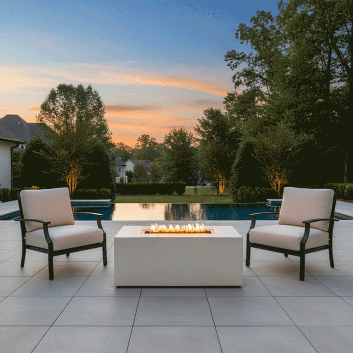 In a suburban yard at sunset, two cushioned armchairs with dark frames stand on a light tile terrace facing a white patio fire pit shaped as a long rectangular fire pit, its narrow opening filled with dark rocks and a line of short flames, set beside a rectangular swimming pool bordered by lawn, hedges, and trees, so the seating and glowing central burner form a straightforward backyard fire pit grouping overlooking the water.