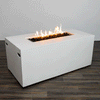 A white rectangular fire pit with smooth vertical sides stands alone on a dark wood floor against a plain light wall, the concrete fire pit showing subtle cutout handles near each end and a long, narrow opening recessed into the top surface, where rough black lava rocks surround a row of bright orange flames rising in uneven peaks, the clean block shape and uncluttered setting emphasizing the piece as a simple modern fire pit.