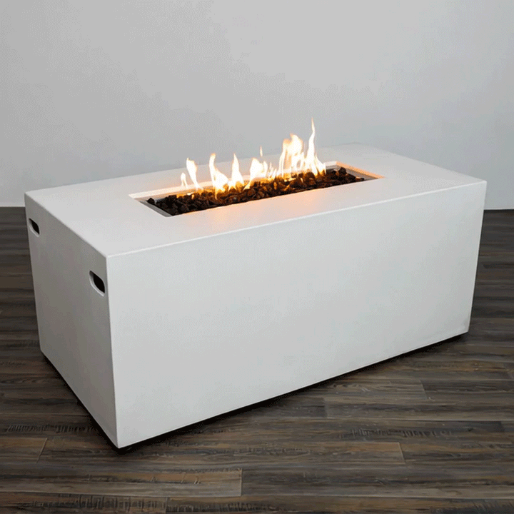 A white rectangular fire pit with smooth vertical sides stands alone on a dark wood floor against a plain light wall, the concrete fire pit showing subtle cutout handles near each end and a long, narrow opening recessed into the top surface, where rough black lava rocks surround a row of bright orange flames rising in uneven peaks, the clean block shape and uncluttered setting emphasizing the piece as a simple modern fire pit.