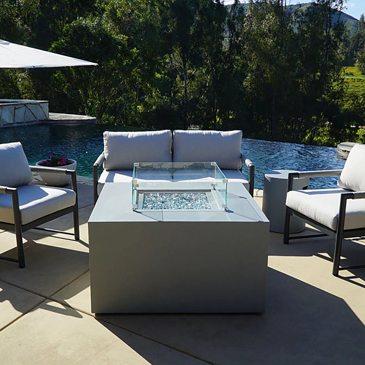 Starfire Designs Beton Square Glass Wind Guard installed on a fire table in a poolside setting. Flames reflect against the tempered glass panels, adding elegance to outdoor lounging areas with modern furniture and lush greenery.