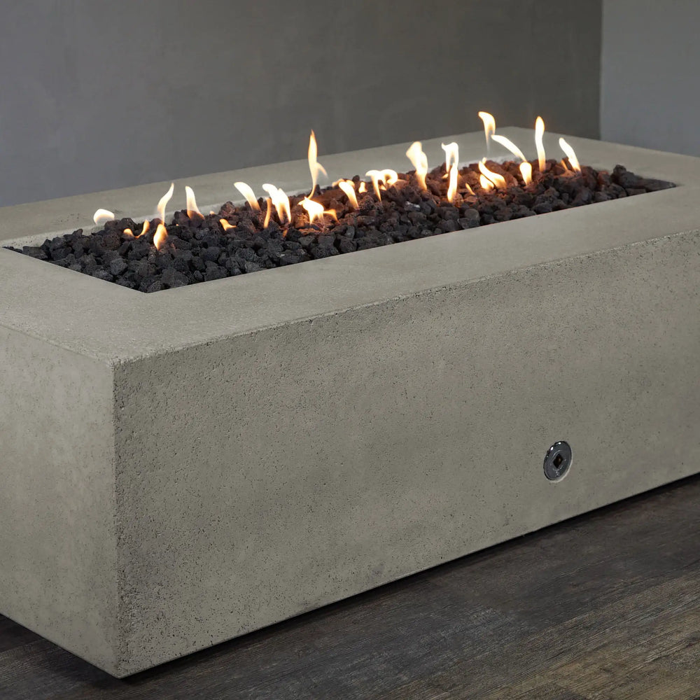 Side-angle view of the Starfire Designs Concrete Rustic 60" x 32" Rectangle Gravity Gas Fire Pit in beige, with flames glowing from the center and lava rocks spread across the burner. The robust concrete structure is perfect for outdoor use, providing both style and functionality in patio and backyard settings