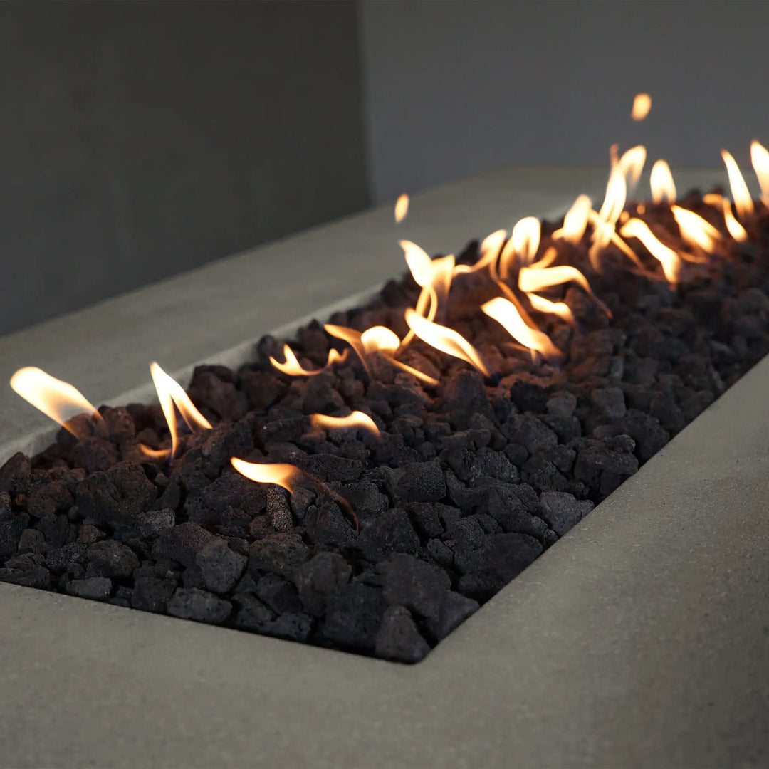 Detailed close-up of black lava rocks inside the Starfire Designs Concrete Rustic 72" x 38" Rectangle Gravity Gas Fire Pit in Beige. The image focuses on the texture of the lava rocks and the steady flames, providing a glimpse into the fire pit's functional and aesthetic appeal.