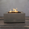 A studio close-up displays a square fire pit in a brown concrete finish, resting on a dark wood floor. Bright yellow flames burn steadily from a bed of black lava rocks, highlighting the earth-tone texture of the concrete fire pit. The visible chrome key valve suggests a manual natural gas fire pit connection, demonstrating the unit's clean design and functionality as a brown fire pit.