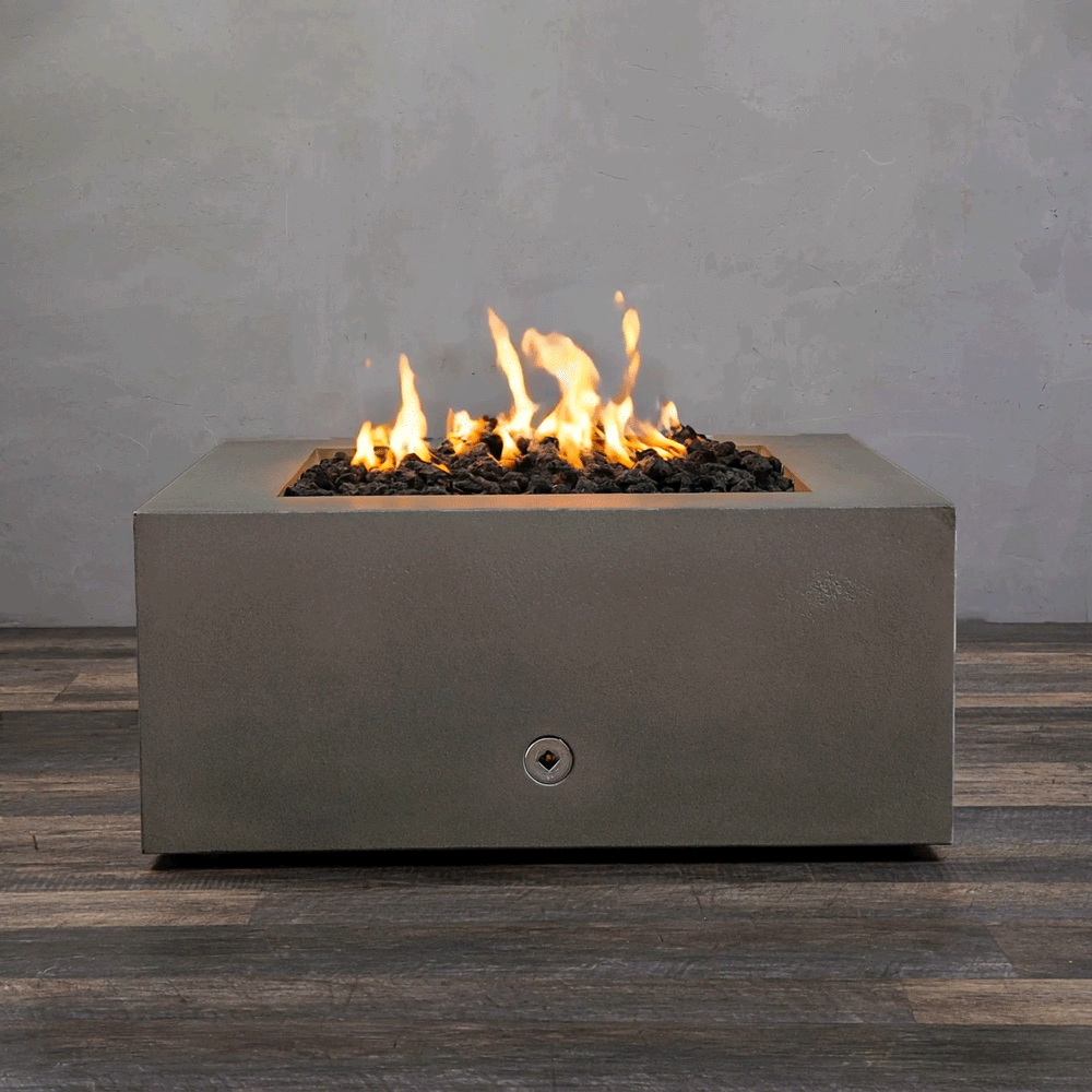 A studio close-up displays a square fire pit in a brown concrete finish, resting on a dark wood floor. Bright yellow flames burn steadily from a bed of black lava rocks, highlighting the earth-tone texture of the concrete fire pit. The visible chrome key valve suggests a manual natural gas fire pit connection, demonstrating the unit's clean design and functionality as a brown fire pit.