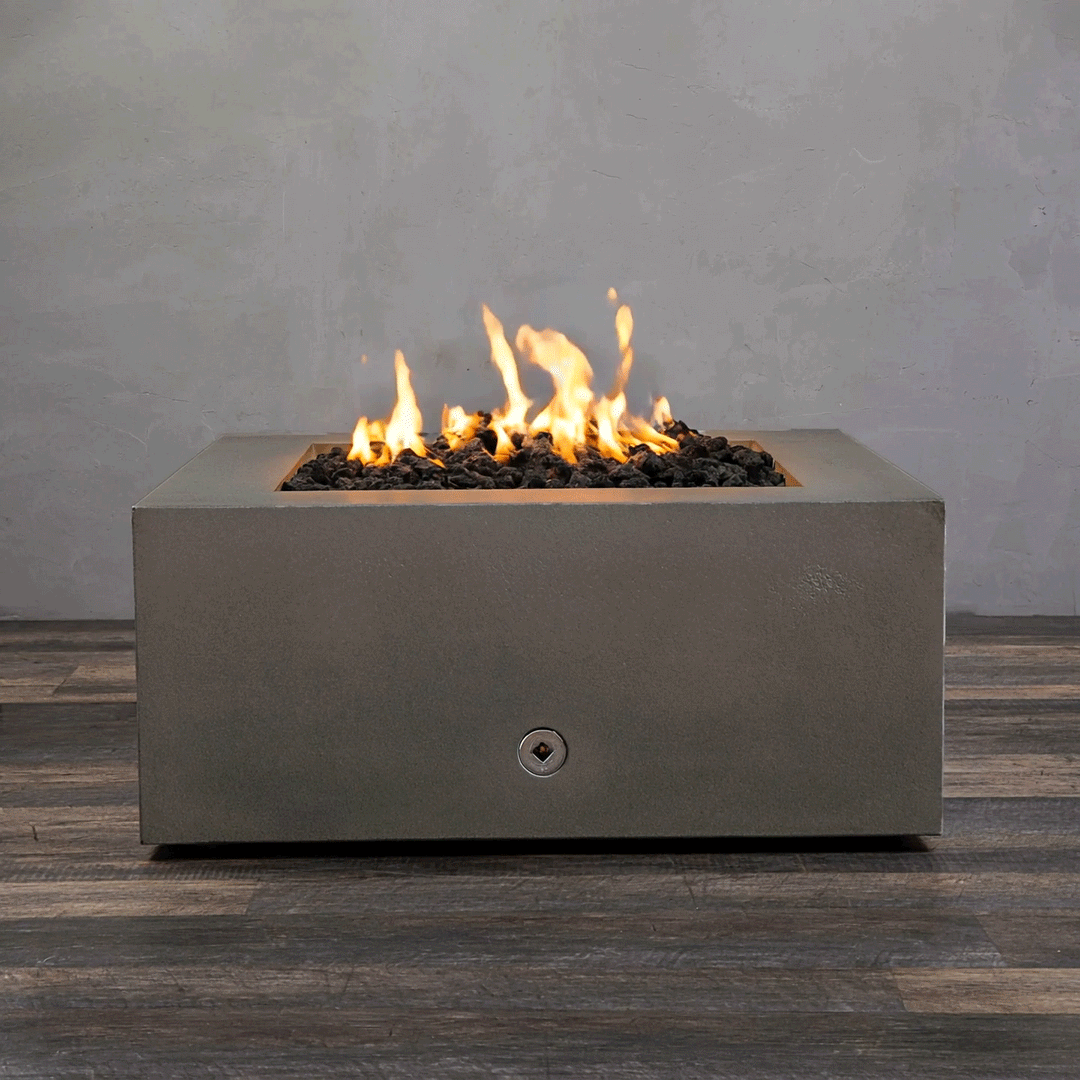 A studio close-up displays a square fire pit in a brown concrete finish, resting on a dark wood floor. Bright yellow flames burn steadily from a bed of black lava rocks, highlighting the earth-tone texture of the concrete fire pit. The visible chrome key valve suggests a manual natural gas fire pit connection, demonstrating the unit's clean design and functionality as a brown fire pit.