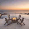 Four wooden Adirondack chairs surround a brown outdoor fire pit on a stone patio overlooking the ocean at sunset. An infinity pool frames the fire feature, which emits a warm glow against the horizon. The setting illustrates a cozy backyard fire pit arrangement, utilizing a propane fire pit to enhance the coastal ambiance.