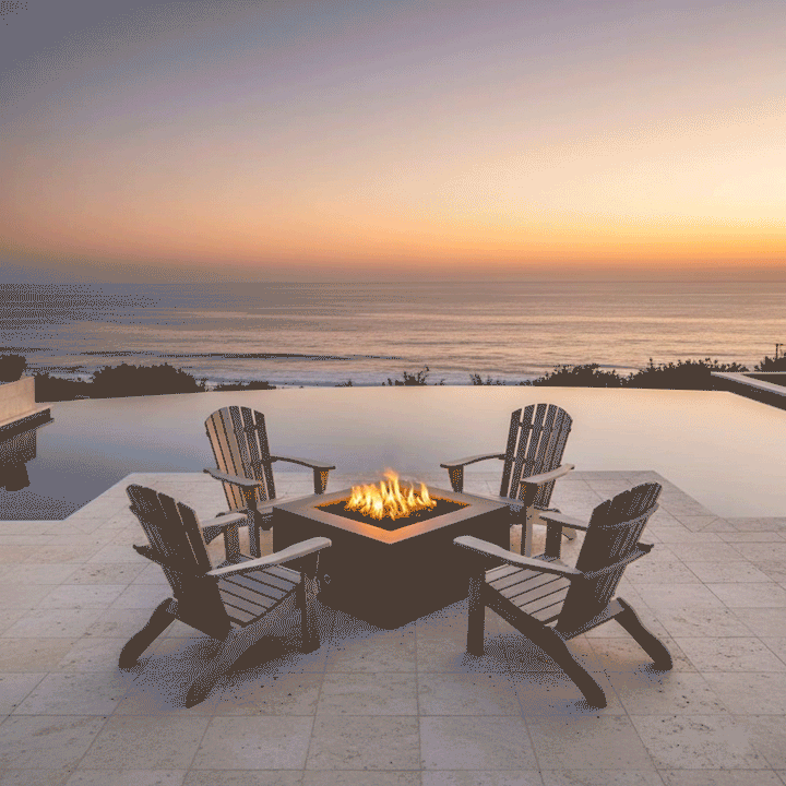 Four wooden Adirondack chairs surround a brown outdoor fire pit on a stone patio overlooking the ocean at sunset. An infinity pool frames the fire feature, which emits a warm glow against the horizon. The setting illustrates a cozy backyard fire pit arrangement, utilizing a propane fire pit to enhance the coastal ambiance.