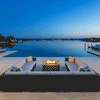 A large U-shaped sectional sofa frames a luxury fire pit in a brown finish, positioned on a waterfront terrace during blue hour. The steady flames provide a warm focal point against the deep blue lake and sky, creating an ideal spot for outdoor entertaining. This family gathering fire pit setup demonstrates the clean, smokeless fire pit operation in a high-end residential environment.