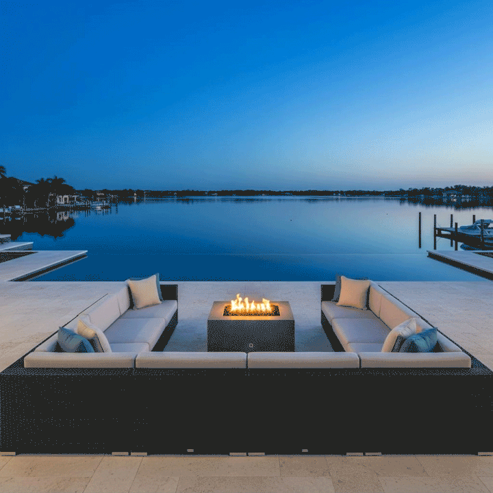 A large U-shaped sectional sofa frames a luxury fire pit in a brown finish, positioned on a waterfront terrace during blue hour. The steady flames provide a warm focal point against the deep blue lake and sky, creating an ideal spot for outdoor entertaining. This family gathering fire pit setup demonstrates the clean, smokeless fire pit operation in a high-end residential environment.