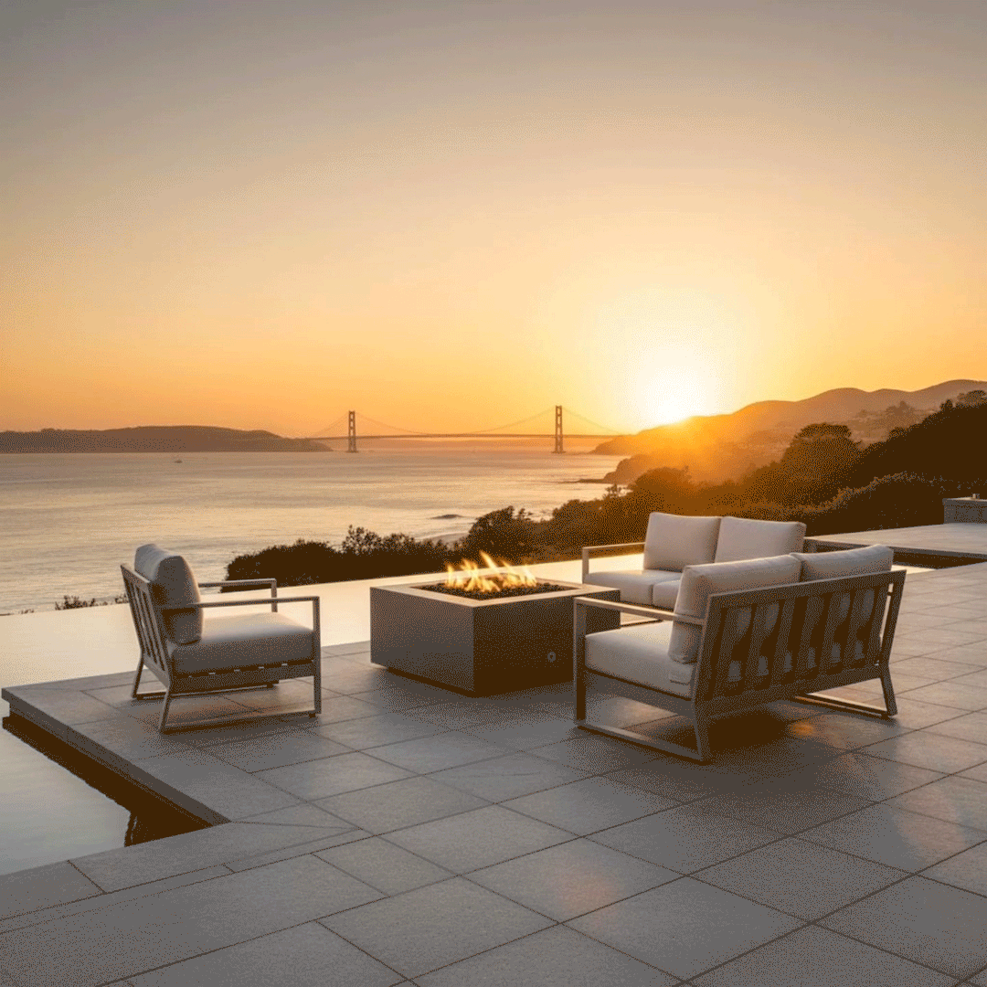 A modern fire pit with a square brown profile sits on a tiled deck overlooking a bay and suspension bridge at sunset. Surrounded by contemporary metal furniture, the patio fire pit anchors the outdoor living space. The amber flames of the gas fire pit blend with the golden hour light, serving as a functional and aesthetic fire table for the scenic overlook.